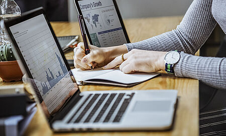 Blick auf einen Schreibtisch mit 2 Laptops und einer Person die etwas notiert.