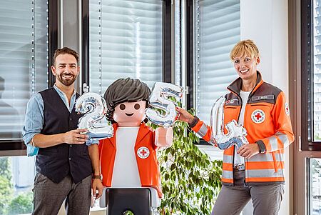 Auf dem Foto sieht man von links: Daniel Matthes von Experts & Talents Dresden, eine fast lebensechte Playmobil-Figur mit braunen Haaren und Rotkreuz-Jacke und Franziska Weiß vom Deutschen Roten Kreuz in Sachsen. Sie alle halten Luftballons hoch, welche die Zahlen 254 zeigen.