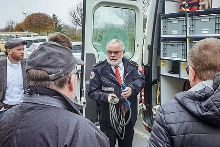 Ein Mitarbeiter des DRK Suchdienstes steht vor einem Einsatzfahrzeug und erklärt dessen Funktion mehreren Zuhörern.