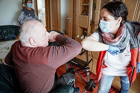 Die Pflegerin und der zu pflegende Mann im Rollstuhl begrüßen sich mit den Ellebogen und tragen einen Mundschutz.