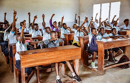 Schüler und Schülerinnen in einem Klassenzimmer in der Schule. Alle melden sich. 