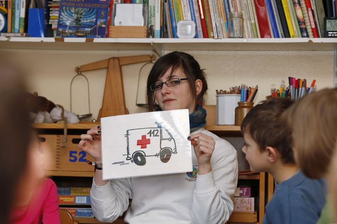 Frau zeigt Kindern ein Schild mit der Notrufnummer 112